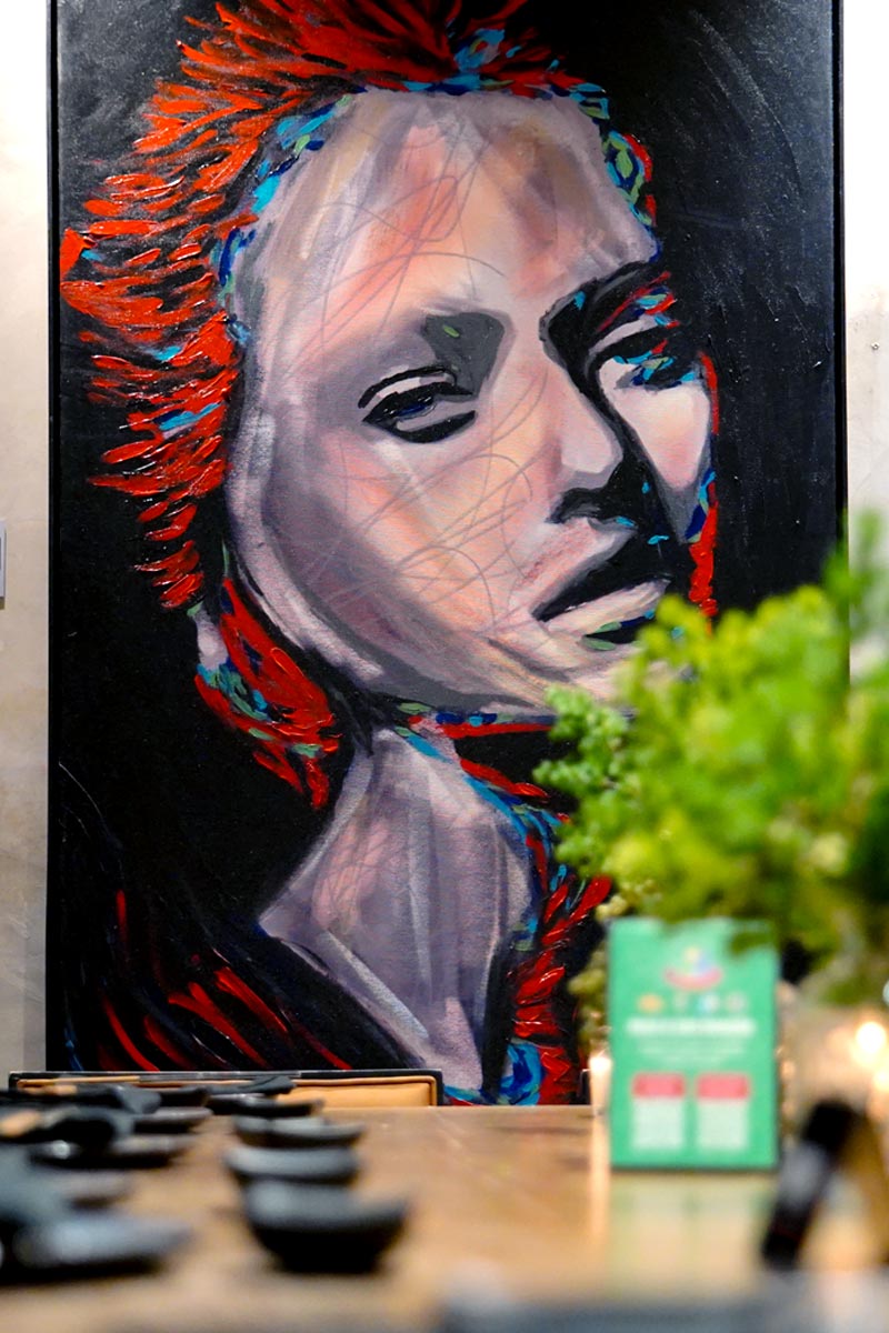 Vivid contemporary painting by Juca Máximo featuring a striking red-haired female figure displayed in a modern restaurant setting.