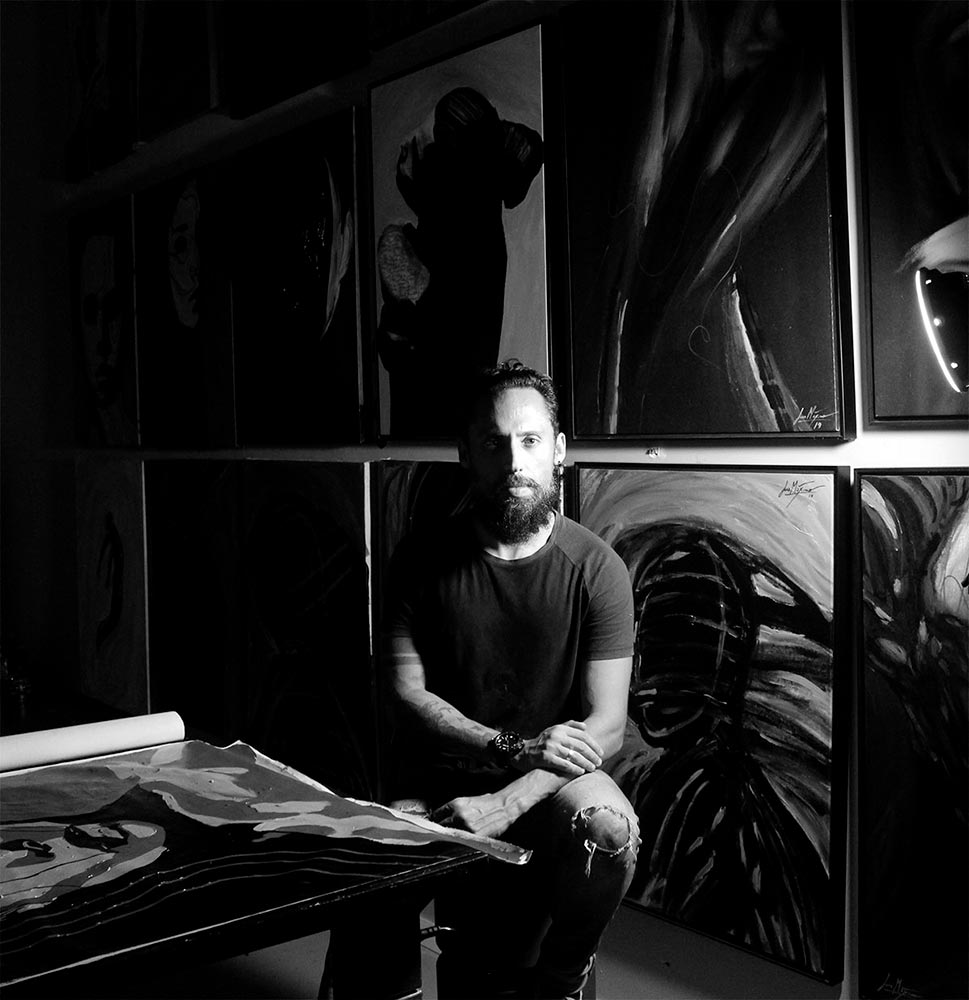 Juca Máximo preparing to paint on a large white canvas in his dimly lit studio, with a focused spotlight and drawings in the background.