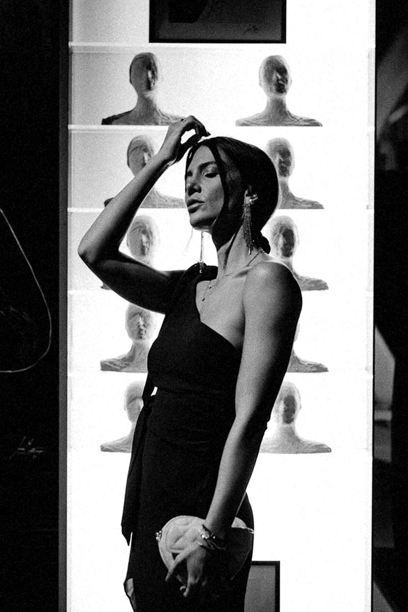 Art model posing in front of illuminated bust sculptures by Juca Maximo at a contemporary gallery exhibition.