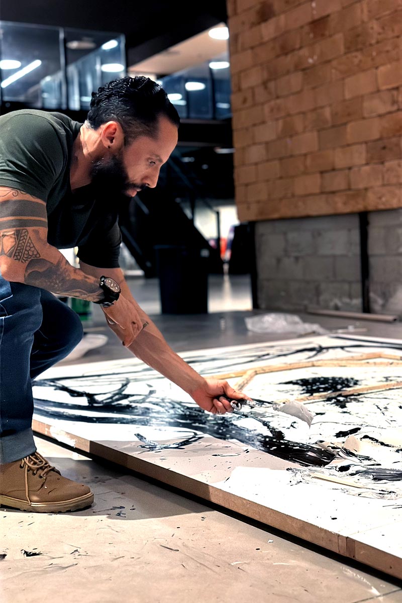Juca Máximo working on a large black and white abstract painting, captured mid-gesture with palette knife during the creative process.
