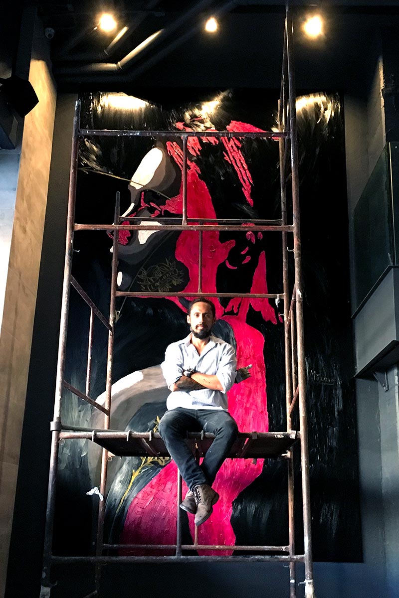 Artist Juca Máximo sitting on scaffolding in front of a monumental pink and black wall panel, portraying strength and creative intensity.