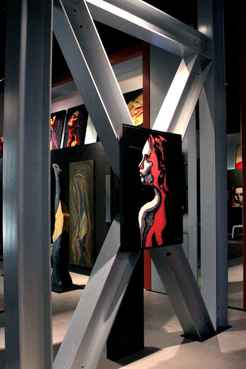 Contemporary emotional painting by Juca Máximo displayed within a steel-structured art exhibition — striking red and black portrait evoking vulnerability and strength.