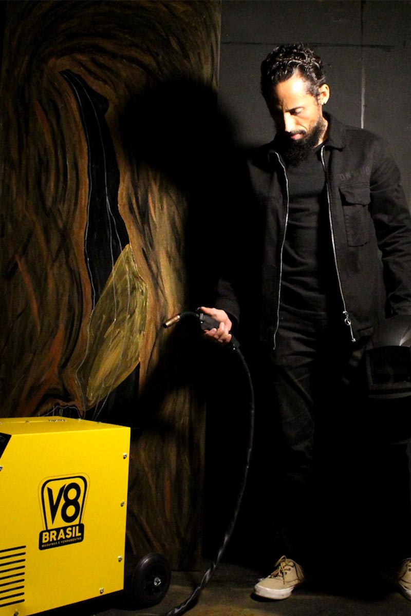 Juca Máximo beside a sculptural panel, holding a welding torch next to a yellow V8 Brasil machine.