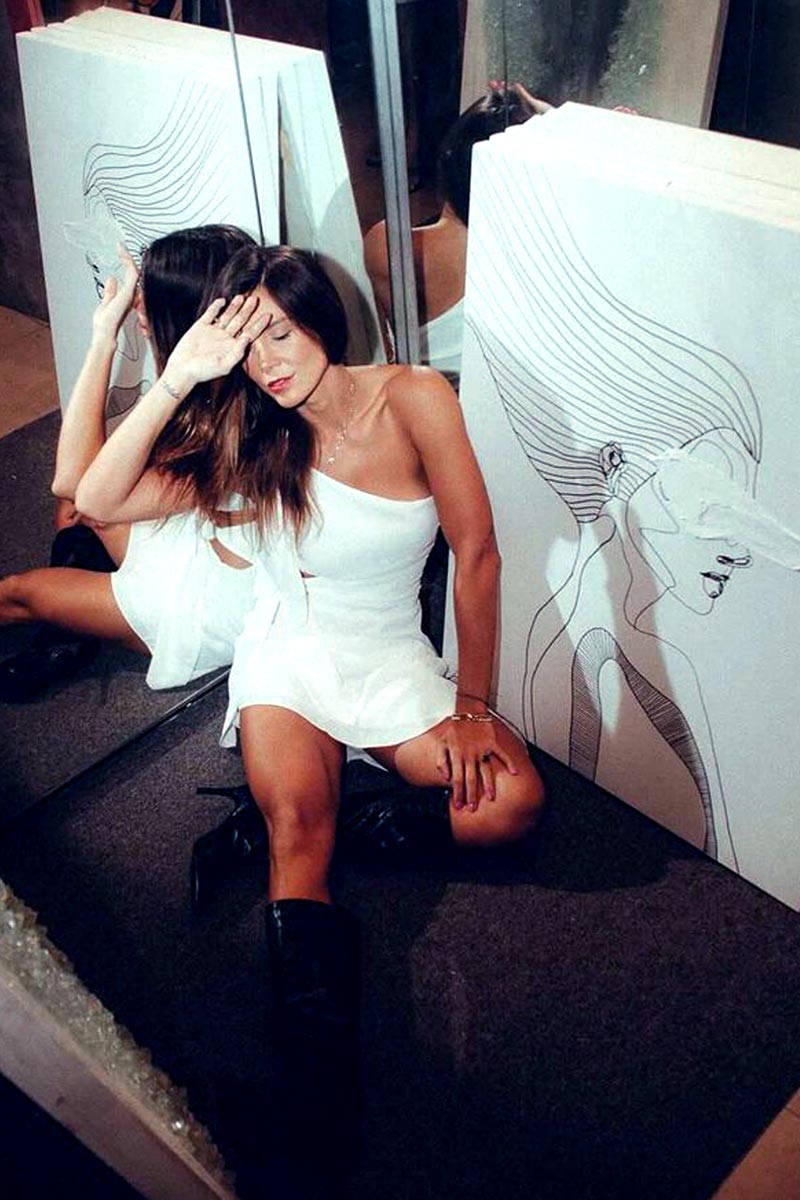 Contemporary art model seated on the floor in front of a large minimalist canvas from the “Traces” series by Juca Máximo, wearing a white dress and black boots, reflecting in a mirror. Emotional connection between the model and the expressive line art portrait enhances the atmosphere of introspection and femininity.