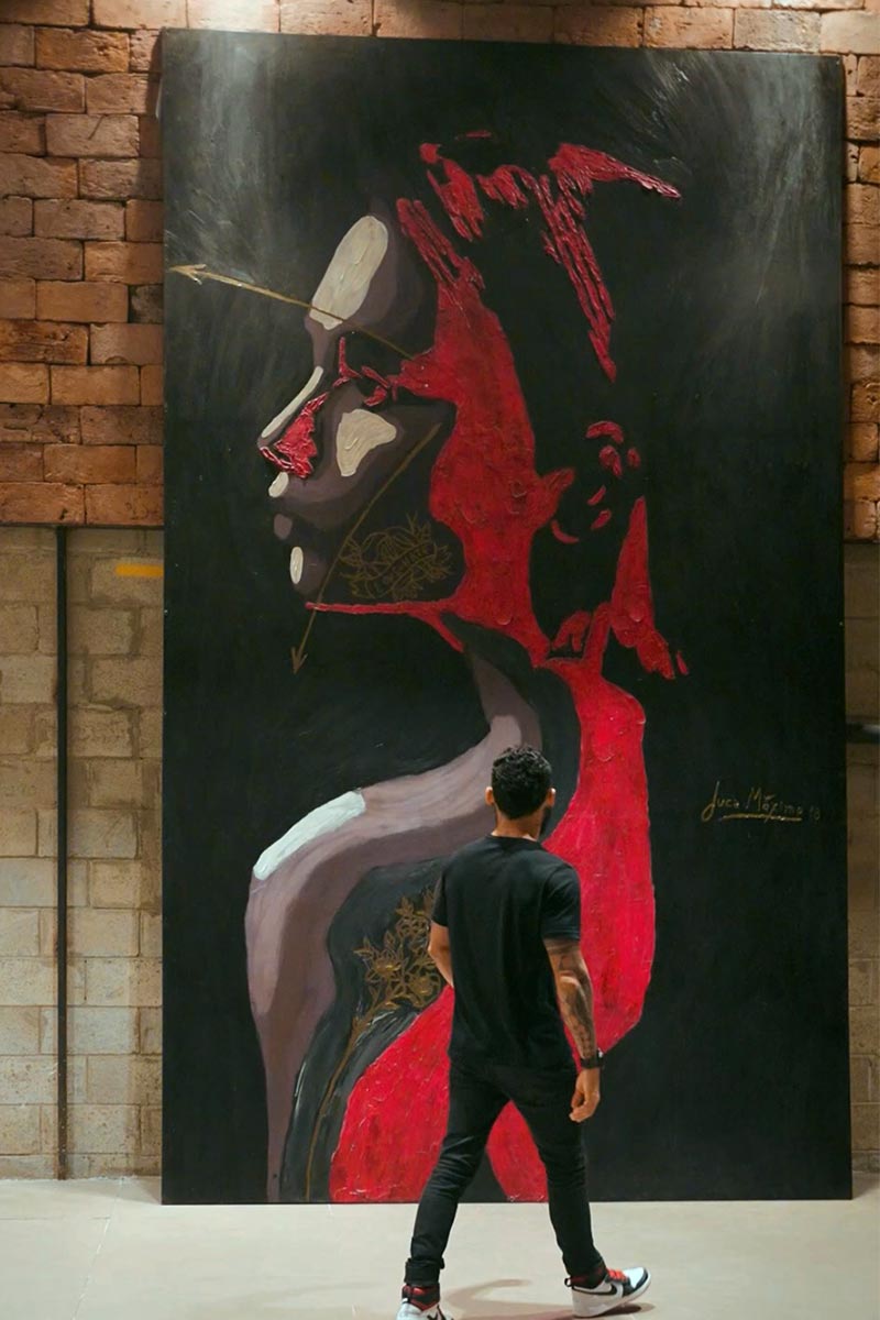 Juca Máximo standing before a monumental red and black emotional portrait artwork — large-scale fine art panel in industrial setting.