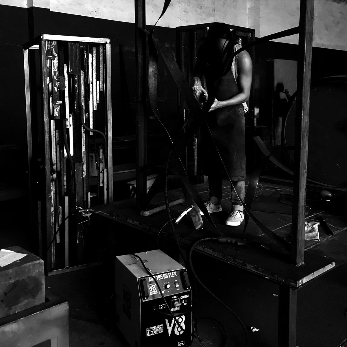Artist welding an abstract metal sculpture structure in a studio space, surrounded by raw materials and tools.