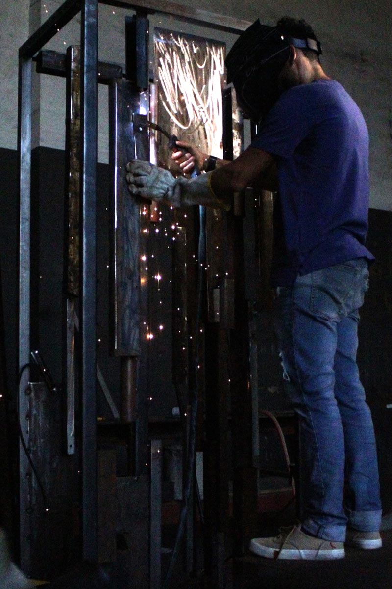 Artist welding a vertical iron frame structure in an industrial studio during the sculpture fabrication process.