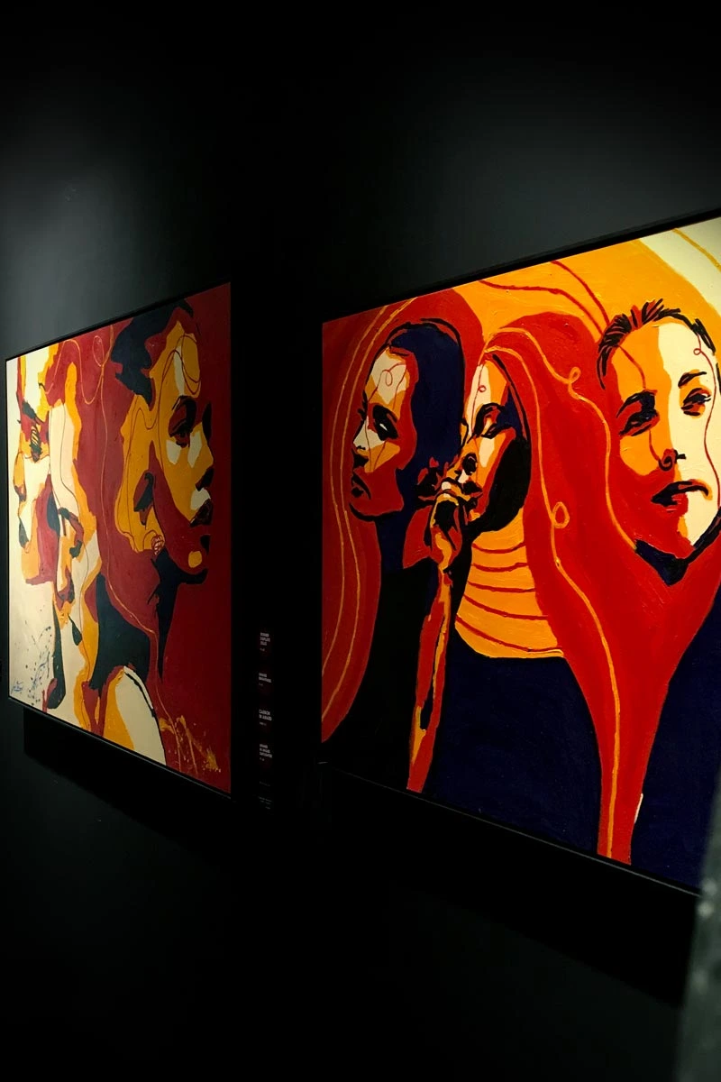 Side view of two paintings from the 'Write Your Story' series in a gallery setting, with dramatic lighting highlighting the bold colors and expressions.