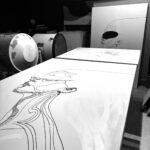 Traces VII in progress resting on a studio table inside Juca Máximo’s atelier — expressive black linework of a female figure on white canvas, surrounded by creative tools and unfinished works in the background.