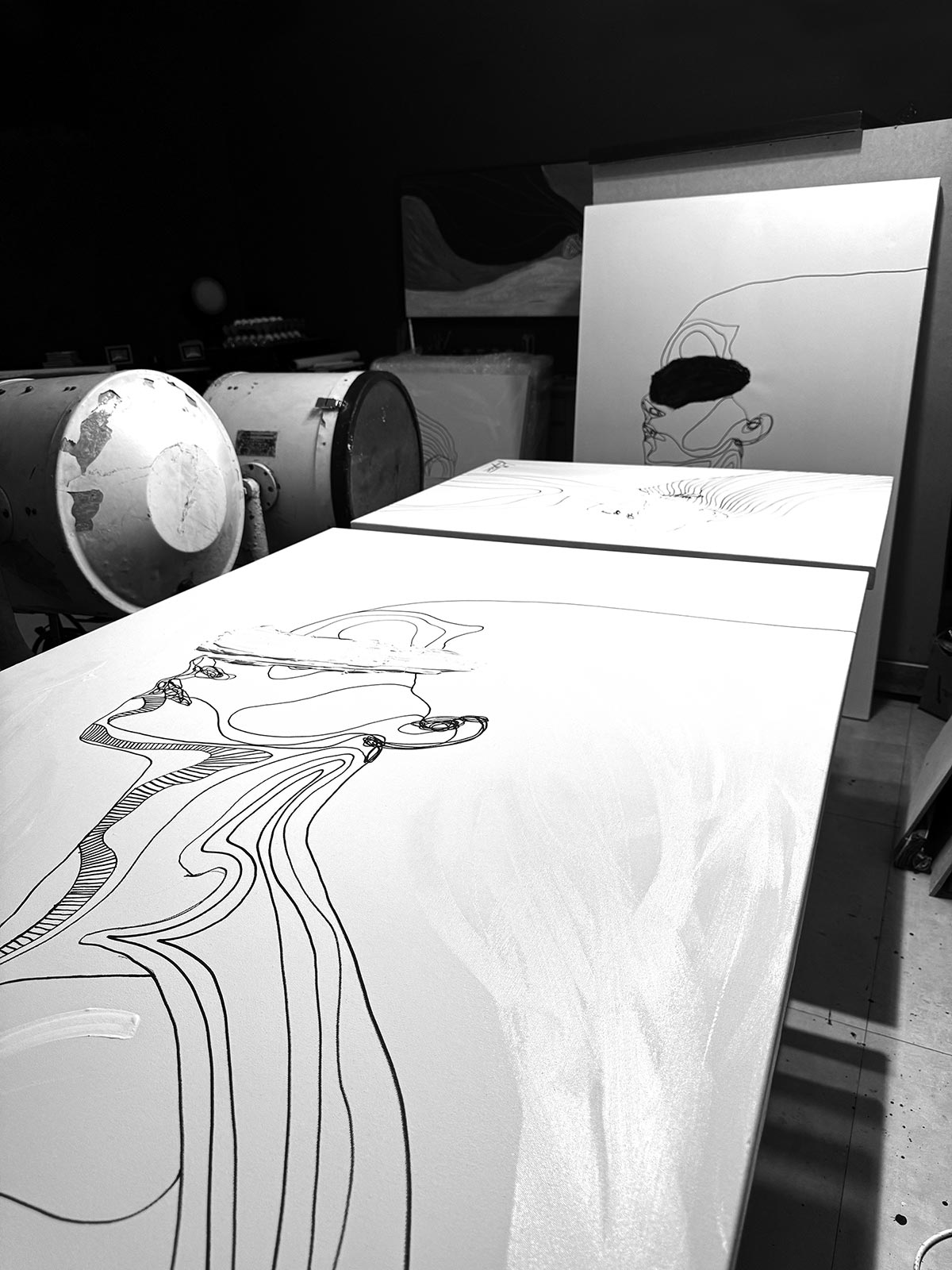 Traces VII in progress resting on a studio table inside Juca Máximo’s atelier — expressive black linework of a female figure on white canvas, surrounded by creative tools and unfinished works in the background.