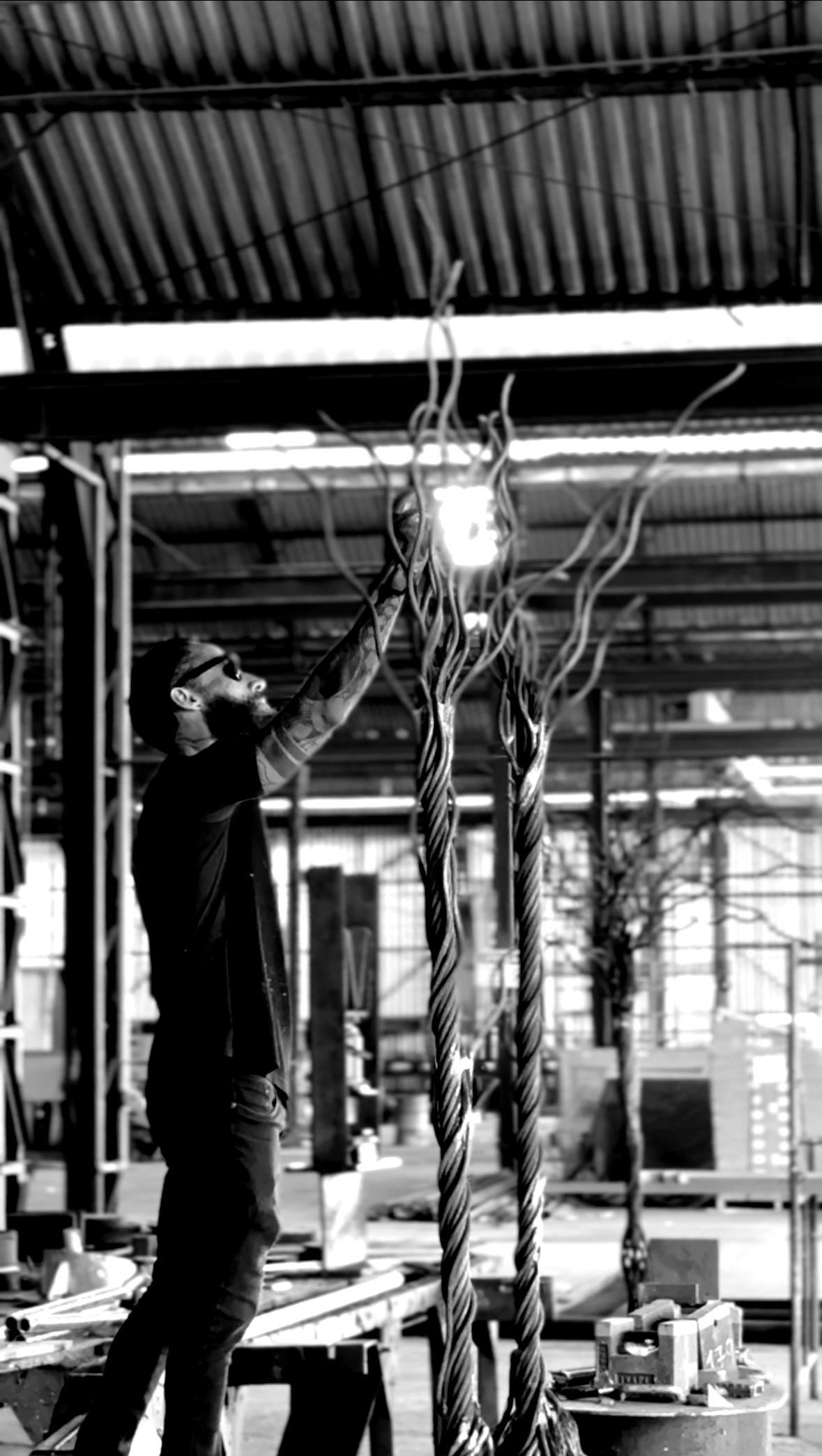 Artist Juca Máximo standing inside an industrial studio, shaping tall vertical sculptures made of twisted steel cables that rise like roots or branches. The black-and-white image highlights his physical gesture, the raw material, and the sculptural process of the installation under construction.