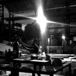 Juca Máximo adjusts a welding gas cylinder inside his industrial art studio at dusk — beams of light and metal shadows reveal the raw poetry of creation, where control meets chaos and silence becomes form.