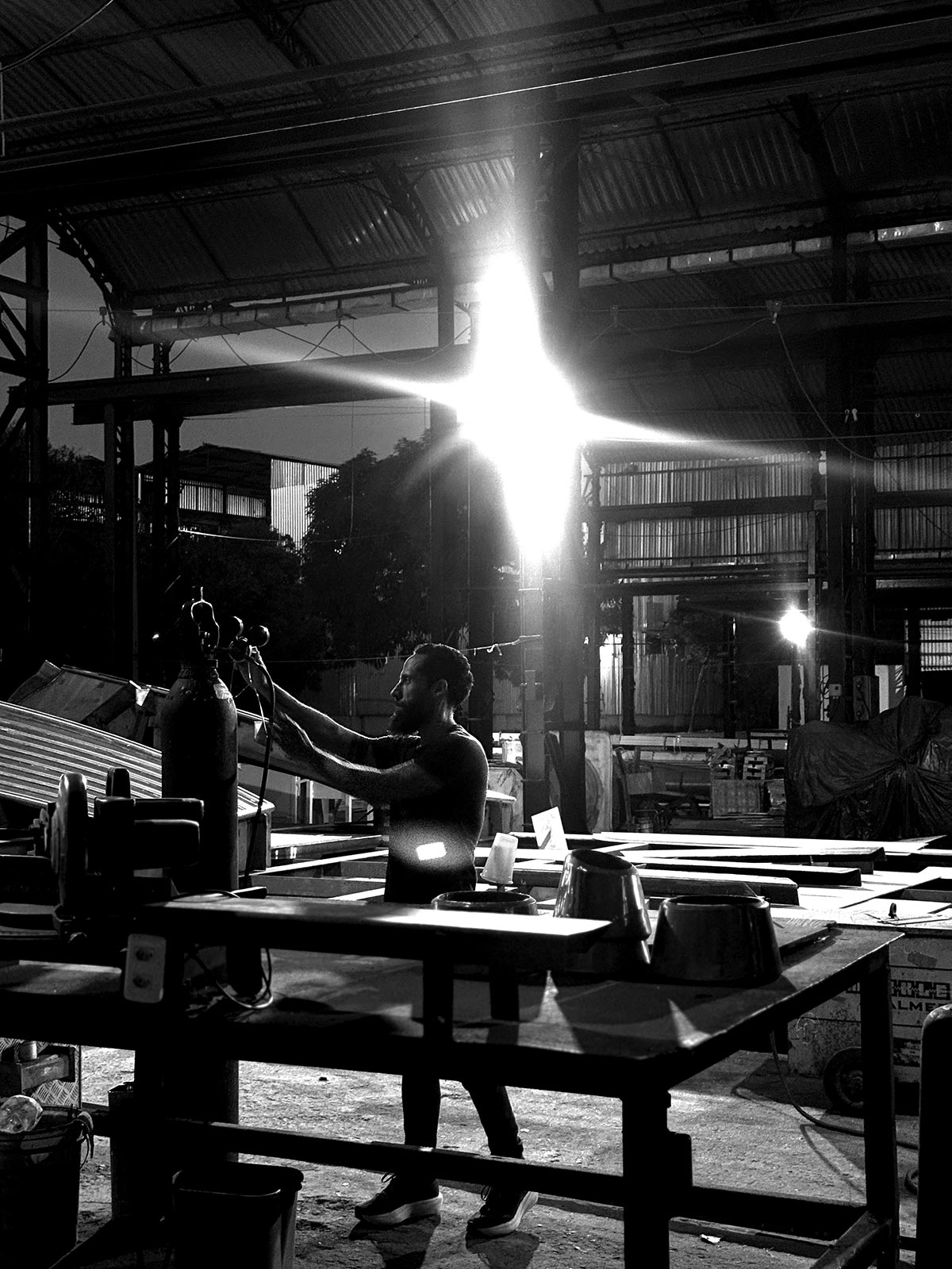 Juca Máximo adjusts a welding gas cylinder inside his industrial art studio at dusk — beams of light and metal shadows reveal the raw poetry of creation, where control meets chaos and silence becomes form.