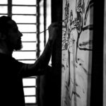 Silhouetted artist painting a large gestural artwork in the studio, holding the brush with fluid precision as light streams through the windows behind; a black-and-white scene capturing creative energy, expressive movement, and the intimate connection between body, gesture, and canvas.
