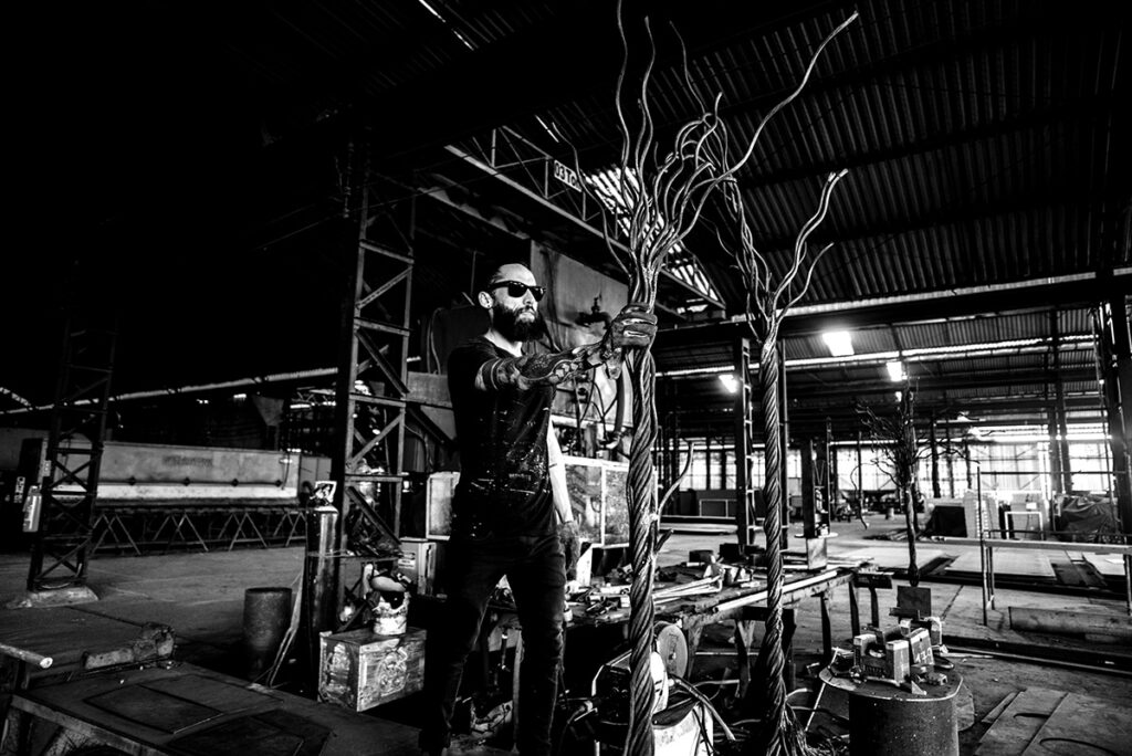Artist Juca Máximo working in his industrial studio, shaping large metallic sculptures called “Swing Trees.” The photo captures the fusion of art and craftsmanship — a poetic scene where strength, gesture, and creation meet amid the raw beauty of steel and light.
