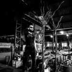 Artist Juca Máximo working in his industrial studio, shaping large metallic sculptures called “Swing Trees.” The photo captures the fusion of art and craftsmanship — a poetic scene where strength, gesture, and creation meet amid the raw beauty of steel and light.