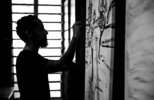 Silhouetted artist painting a large gestural artwork in the studio, holding the brush with fluid precision as light streams through the windows behind; a black-and-white scene capturing creative energy, expressive movement, and the intimate connection between body, gesture, and canvas.