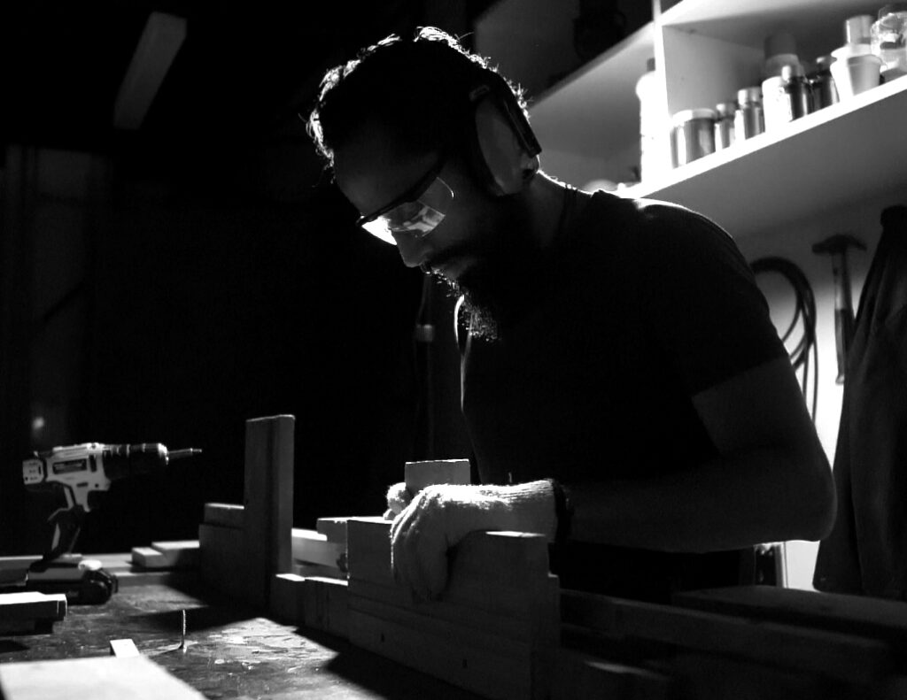 Juca Máximo in his studio working with wood under soft directional light, wearing safety glasses and headphones, shaping a wooden installation surrounded by tools and shelves — a poetic black and white portrait of the artist’s deep sensory connection with the material at Juca Máximo Studio.