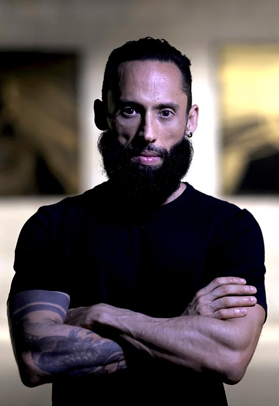 Portrait of artist Juca Máximo standing with arms crossed in his studio, with large contemporary abstract paintings blurred in the background. Strong lighting highlights his face and presence, conveying authority and artistic identity.