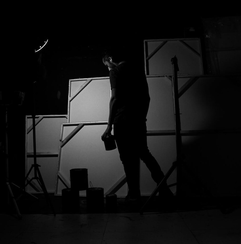 Man standing in a dim studio holding a paint can in front of multiple blank canvases leaning against the wall, with soft side lighting and studio equipment around, representing the beginning of an artistic creation process.