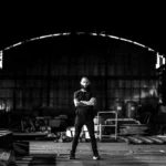 Artista visual em pé no centro de um galpão industrial vazio, com os braços cruzados, cercado por estruturas metálicas, aço e materiais brutos, em um espaço amplo e escuro que remete a força, escala e processo criativo. Contemporary visual artist standing at the center of an empty industrial warehouse, arms crossed, surrounded by metal structures, steel and raw materials, in a large and dark space that evokes strength, scale and the artistic process.