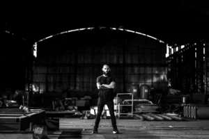 Artista visual em pé no centro de um galpão industrial vazio, com os braços cruzados, cercado por estruturas metálicas, aço e materiais brutos, em um espaço amplo e escuro que remete a força, escala e processo criativo. Contemporary visual artist standing at the center of an empty industrial warehouse, arms crossed, surrounded by metal structures, steel and raw materials, in a large and dark space that evokes strength, scale and the artistic process.