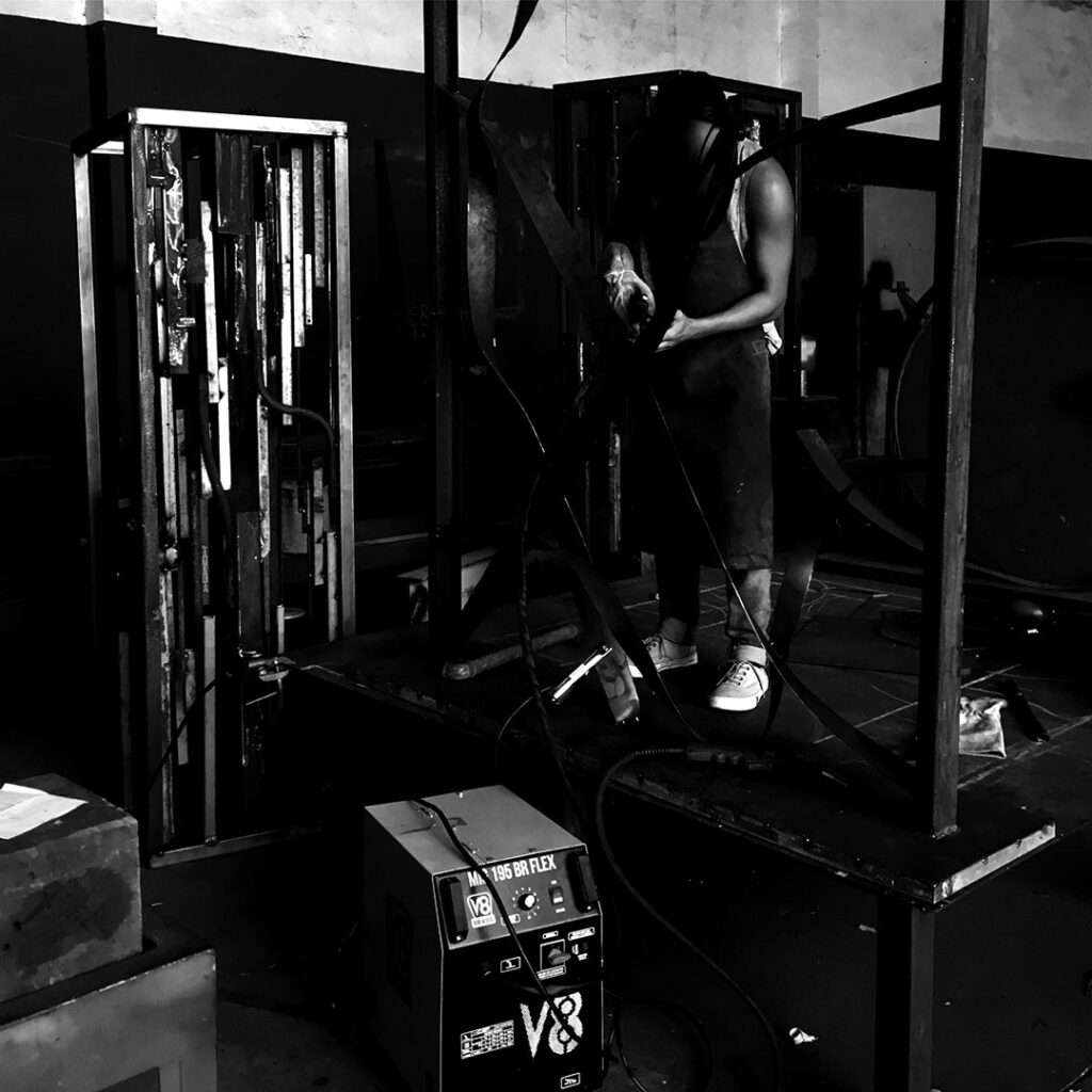 Artista soldando escultura em ferro no estúdio, utilizando máquina de solda em estrutura metálica da série Energy, explorando tensão, fogo e construção física da obra em processo. Artist welding an iron sculpture in the studio, using a welding machine on a metal structure from the Energy series, exploring tension, fire, and the physical construction of the artwork in process.