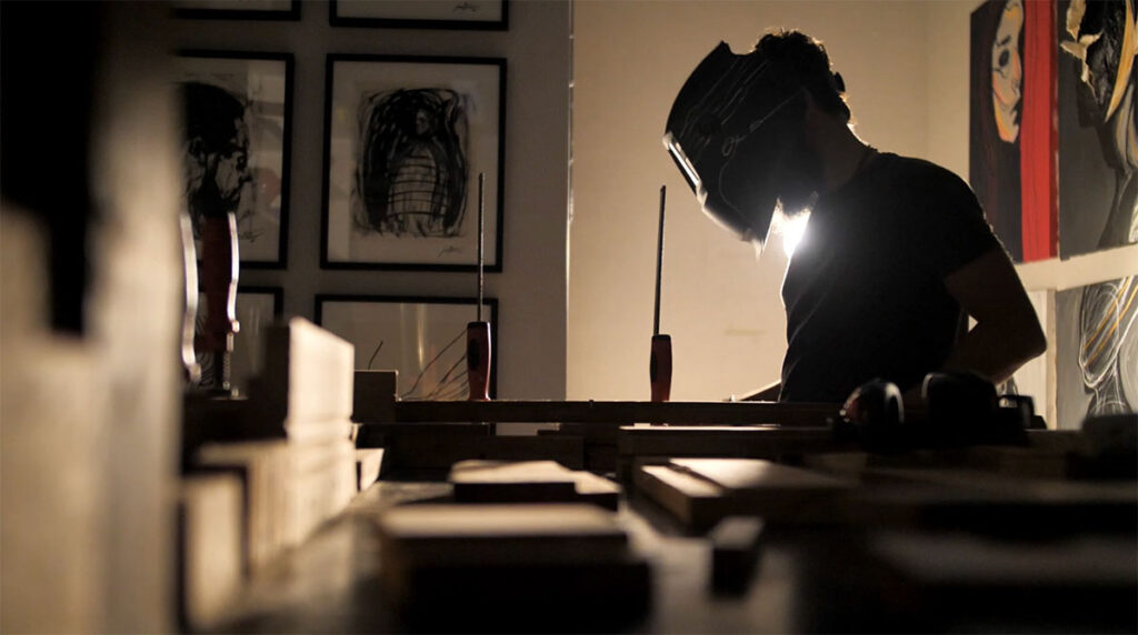 Brazilian contemporary artist Juca Máximo working in his studio, welding a sculpture structure surrounded by figurative paintings and artworks in the background.