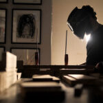 Brazilian contemporary artist Juca Máximo working in his studio, welding a sculpture structure surrounded by figurative paintings and artworks in the background.