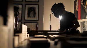 Brazilian contemporary artist Juca Máximo working in his studio, welding a sculpture structure surrounded by figurative paintings and artworks in the background.
