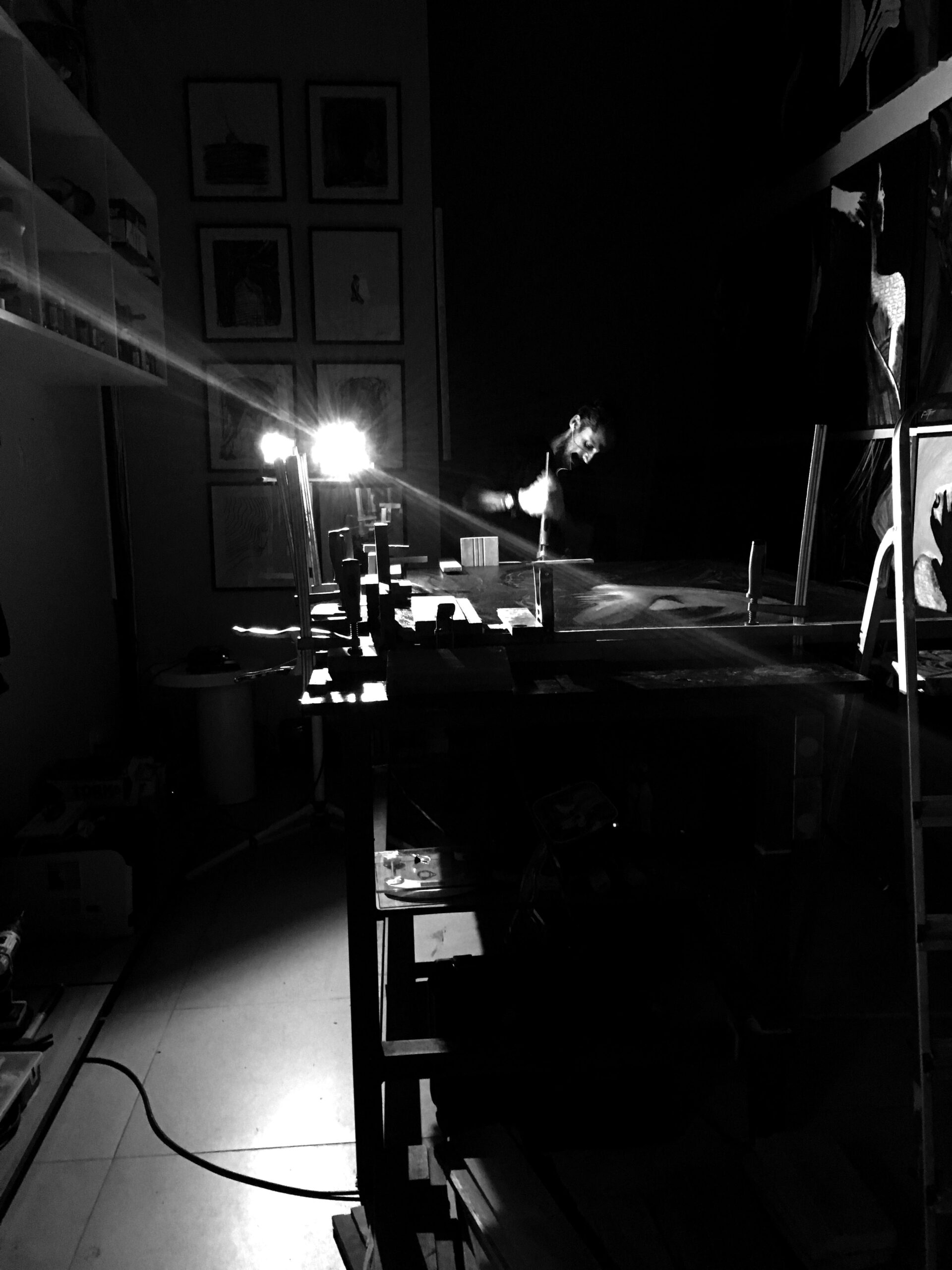 Artist working inside a dimly lit contemporary studio during the creation process of a large-scale abstract artwork. Warm directional lighting illuminates the artist’s gesture and workspace while deep shadows create a cinematic atmosphere. The studio contains wooden structures, clamps, tools, framed monochromatic drawings, and large expressive paintings on dark walls. The image conveys immersion, intensity, solitude, and the physical relationship between body, gesture, and artistic creation. Contemporary fine art studio environment with minimalist industrial aesthetics and emotional ambience.