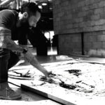 Juca Máximo ajoelhado sobre o chão de seu estúdio enquanto trabalha diretamente sobre uma grande pintura em preto e branco. O artista utiliza uma espátula para espalhar tinta sobre a superfície da obra, cercado por um ambiente industrial e contemporâneo. A fotografia em preto e branco destaca o gesto manual, o processo criativo e a relação física entre artista e obra em desenvolvimento. Juca Máximo kneels on the floor of his studio while working directly on a large black-and-white painting. Using a palette knife, the artist spreads paint across the surface of the artwork within a contemporary industrial environment. The black-and-white photograph emphasizes the physical gesture, creative process, and the connection between artist and artwork during its creation.
