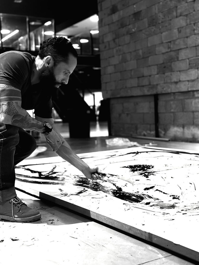 Juca Máximo ajoelhado sobre o chão de seu estúdio enquanto trabalha diretamente sobre uma grande pintura em preto e branco. O artista utiliza uma espátula para espalhar tinta sobre a superfície da obra, cercado por um ambiente industrial e contemporâneo. A fotografia em preto e branco destaca o gesto manual, o processo criativo e a relação física entre artista e obra em desenvolvimento. Juca Máximo kneels on the floor of his studio while working directly on a large black-and-white painting. Using a palette knife, the artist spreads paint across the surface of the artwork within a contemporary industrial environment. The black-and-white photograph emphasizes the physical gesture, creative process, and the connection between artist and artwork during its creation.