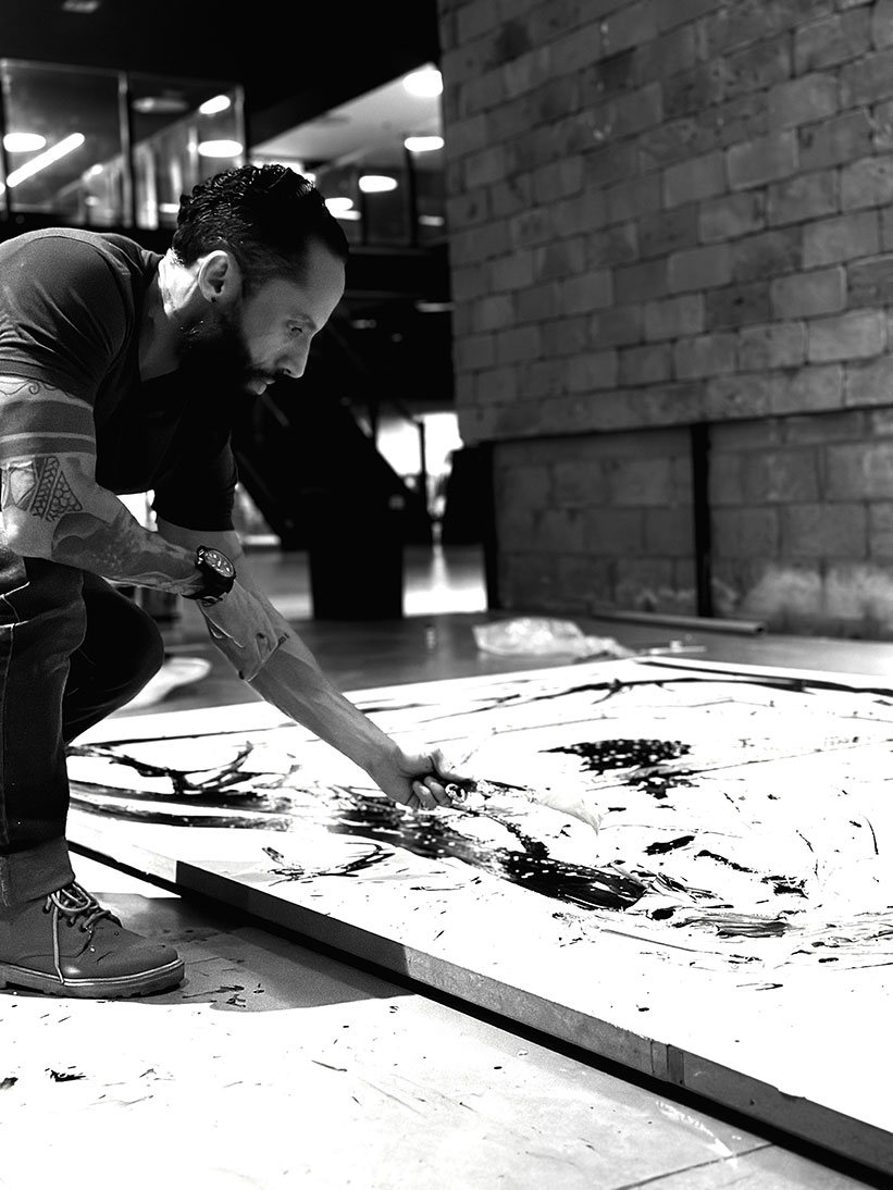 Juca Máximo ajoelhado sobre o chão de seu estúdio enquanto trabalha diretamente sobre uma grande pintura em preto e branco. O artista utiliza uma espátula para espalhar tinta sobre a superfície da obra, cercado por um ambiente industrial e contemporâneo. A fotografia em preto e branco destaca o gesto manual, o processo criativo e a relação física entre artista e obra em desenvolvimento. Juca Máximo kneels on the floor of his studio while working directly on a large black-and-white painting. Using a palette knife, the artist spreads paint across the surface of the artwork within a contemporary industrial environment. The black-and-white photograph emphasizes the physical gesture, creative process, and the connection between artist and artwork during its creation.